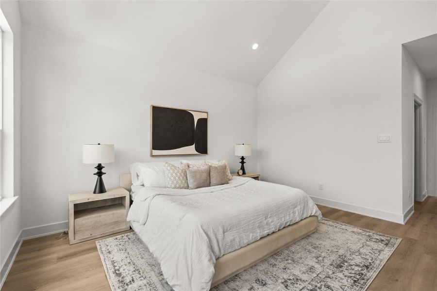 This view of the primary suite shows off its balanced layout, high vaulted ceiling, and smooth white walls. Light oak flooring runs throughout the space, while black bedside lamps and an abstract art piece add a clean, modern touch to the room.