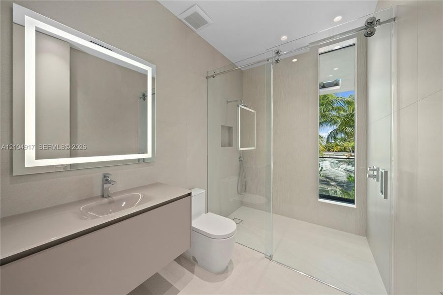 Floating vanity with integrated sink and chrome faucet, illuminated mirrored medicine cabinet, glass-enclosed shower, wall-mounted shower fixtures, and a vertical window