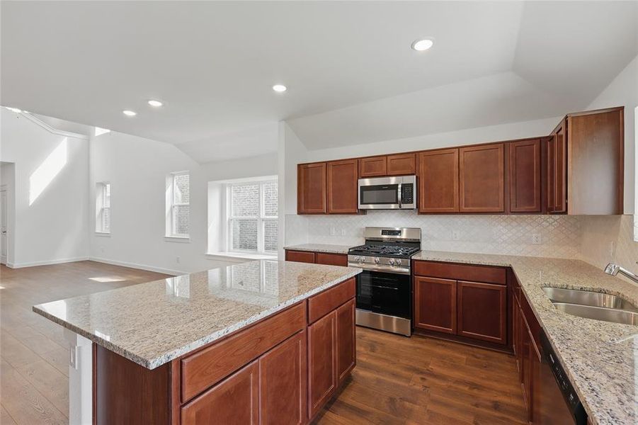 Kitchen with tasteful backsplash, appliances with stainless steel finishes, light stone counters, dark wood-style floors, and vaulted ceiling