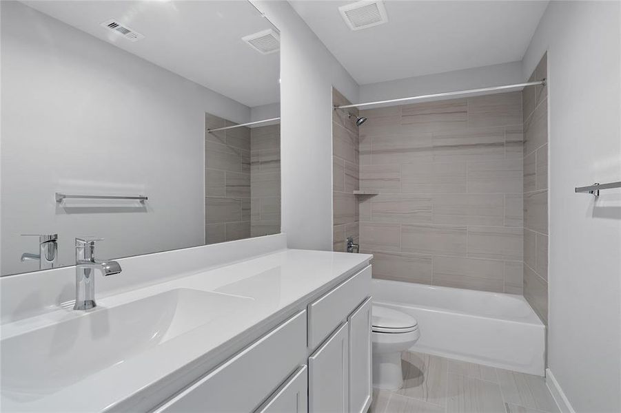 Secondary bath features tile flooring, bath/shower combo with tile surround, white stained wood cabinets, beautiful light countertops, mirror, dark, sleek fixtures and modern finishes.