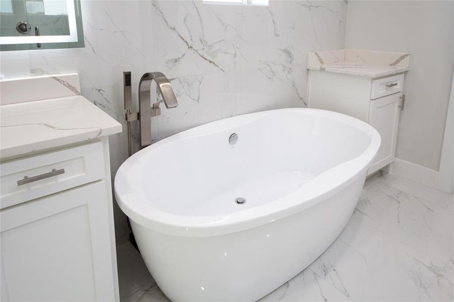 Bathroom featuring light marble finish floors, vanity, a freestanding tub, and stone wall