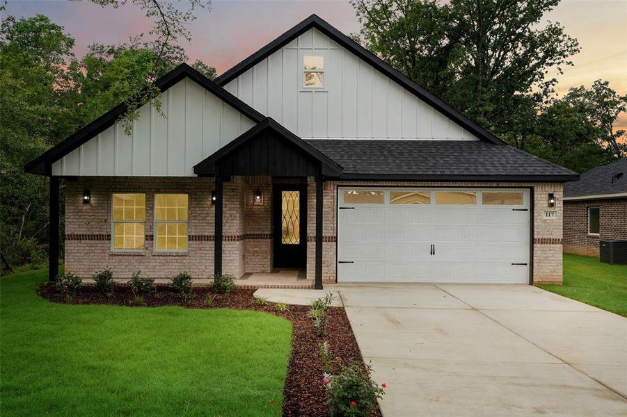 Exterior featuring a brick facade, white board and batten siding, dark shingle roof, attached garage with decorative hardware, and a landscaped front yard with a concrete driveway Exterior featuring a brick facade, white board and batten siding, dark shingle roof, attached garage with decorative hardware, and a landscaped front yard with a concrete driveway