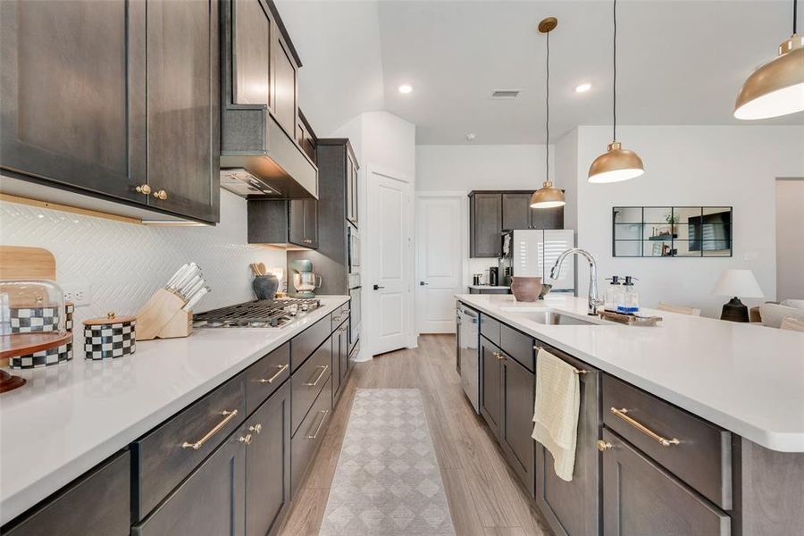 Modern kitchen featuring a large island with sink, white countertops, dark wood-finish cabinetry with gold-tone hardware, and a gas cooktop with a white tile backsplash