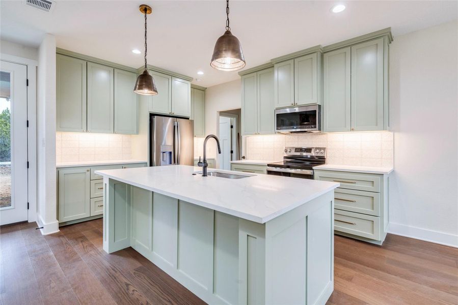 Kitchen with appliances with stainless steel finishes, green cabinetry, light stone counters, pendant lighting, and a center island with sink