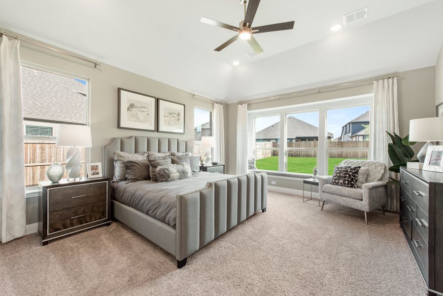 Master bedroom with gray upholstered bed, carpet flooring, ceiling fan, and large windows with backyard view