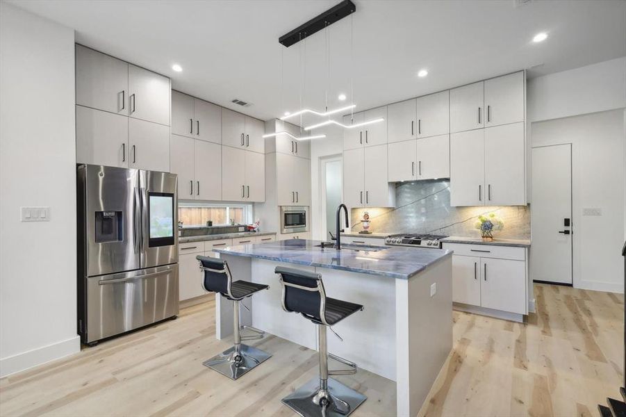 Kitchen with light hardwood / wood-style floors, appliances with stainless steel finishes, hanging light fixtures, and an island with sink