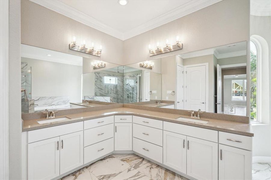 This is a spacious, well-lit bathroom featuring dual vanities with ample storage, elegant marble flooring, and a sleek modern design. Large mirrors enhance the sense of space, and chic light fixtures provide a warm ambiance.