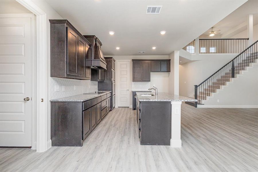 Kitchen with black electric stovetop, ceiling fan, light wood-type flooring, light stone countertops, and an island with sink