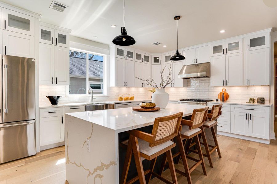 Kitchen featuring white cabinetry, a large kitchen island with a waterfall edge countertop, light hardwood floors, and stainless steel appliances Kitchen featuring white cabinetry, a large kitchen island with a waterfall edge countertop, light hardwood floors, and stainless steel appliances