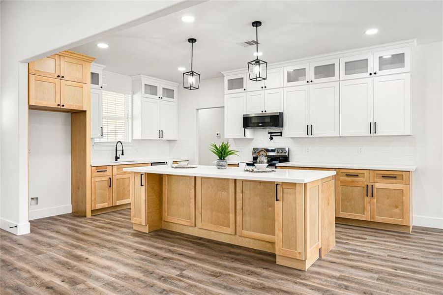 A modern kitchen with a spacious layout features a central island with light wood cabinetry and white countertops. It has overhead pendant lights, white upper cabinets with glass inserts, and stainless steel appliances. The flooring is a warm wood tone, enhancing the bright and airy feel.