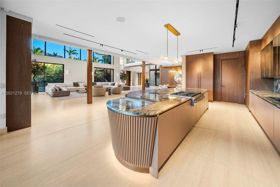 Designer kitchen with curved fluted island, book-matched stone countertop, gas cooktop, brass linear pendant, full-height walnut cabinetry, dramatic stone backsplash with smoked glass uppers, wine refrigerator, and open view to double-height great room and waterfront beyond.