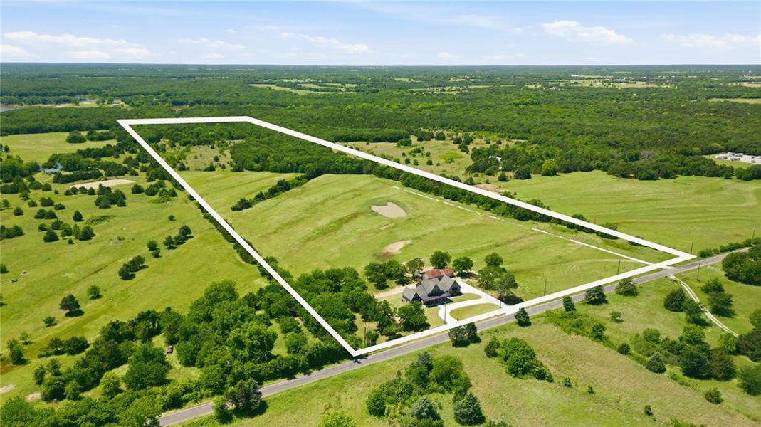 Expansive rural property featuring a primary residence with a dark roof and light-colored exterior, a curved asphalt driveway, and surrounding mature trees
