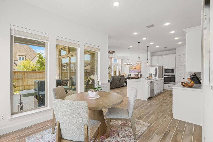 This photo showcases a bright, open-concept breakfast room, kitchen, family room and dining area. The wood-look tile flooring brings continuity throughout the house.