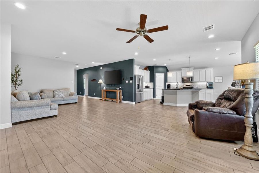 Spacious open-concept living area featuring wood-look tile flooring, recessed lighting, a ceiling fan, and a kitchen with white cabinetry and stainless steel appliances