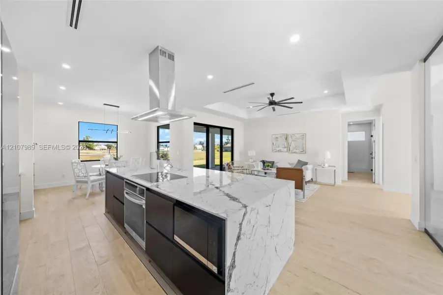 Kitchen featuring a large waterfall-edge island with stone-finish countertops, stainless steel chimney-style range hood, recessed lighting, and wood-finish flooring Kitchen featuring a large waterfall-edge island with stone-finish countertops, stainless steel chimney-style range hood, recessed lighting, and wood-finish flooring