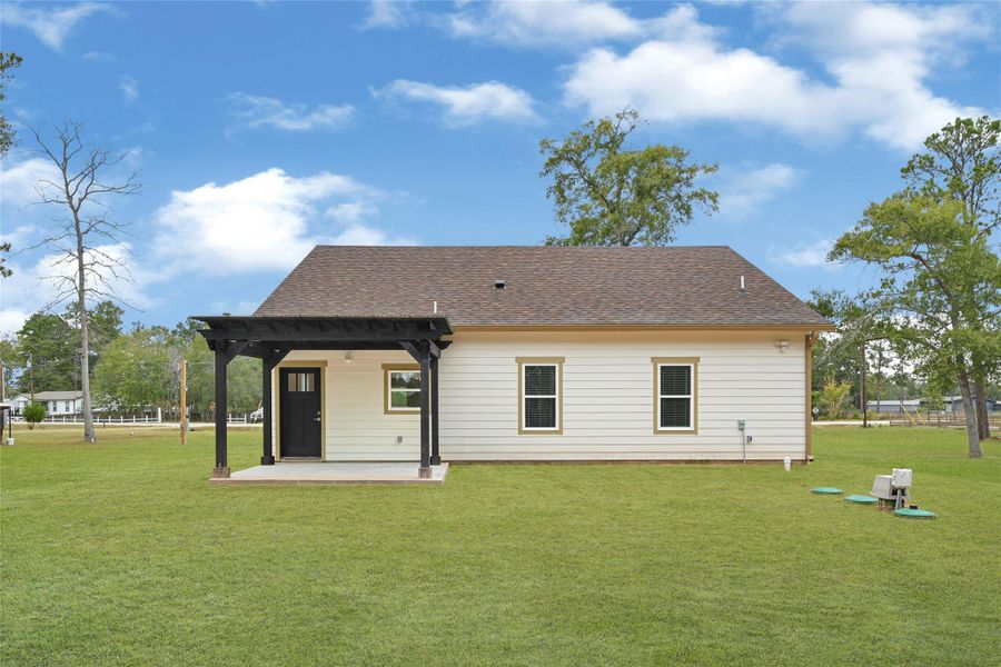This photo shows the rear view of a single-story house featuring a light-colored exterior with a dark pergola over a small patio. It sits on a spacious grassy lot with a few trees, offering a serene and open outdoor space. This photo shows the rear view of a single-story house featuring a light-colored exterior with a dark pergola over a small patio. It sits on a spacious grassy lot with a few trees, offering a serene and open outdoor space.