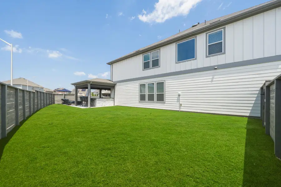 Expansive backyard featuring a lush turf green lawn, covered patio structure with built-in stone counter, and wood-finish perimeter fencing
