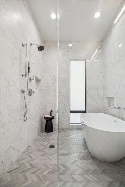 Spa-inspired primary bath wrapped in floor-to-ceiling marble-look tile with a frameless glass walk-in shower and elegant freestanding soaking tub. Chevron-patterned flooring adds visual texture while recessed lighting enhances the bright, airy ambiance. Clean architectural lines, built-in wall niches, and a sleek window panel create a serene retreat designed for relaxation and elevated everyday living.