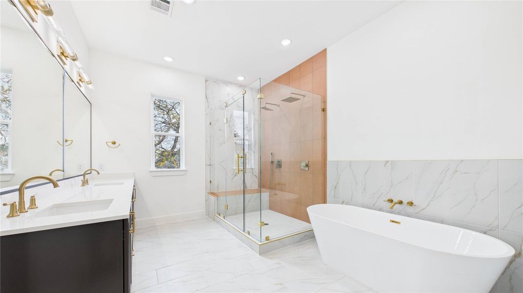 The primary bathroom features a spacious walk-in glass shower with two rainheads and a spray wand, a freestanding bathtub with gold fixtures, and a double vanity with elegant lighting. The room is bright and sleek.