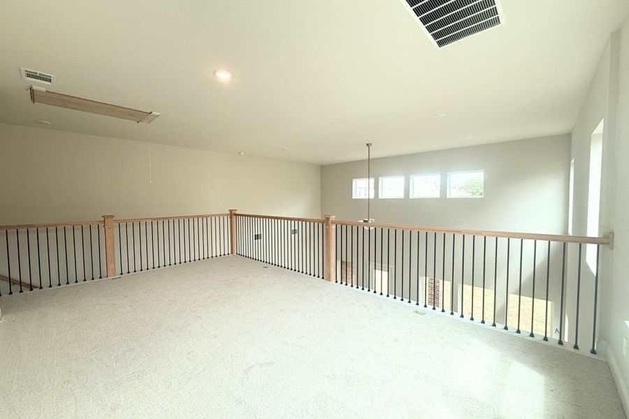 Spacious loft area featuring light-toned carpeting, a wood-finish railing with black iron balusters, and multiple windows providing natural light Spacious loft area featuring light-toned carpeting, a wood-finish railing with black iron balusters, and multiple windows providing natural light