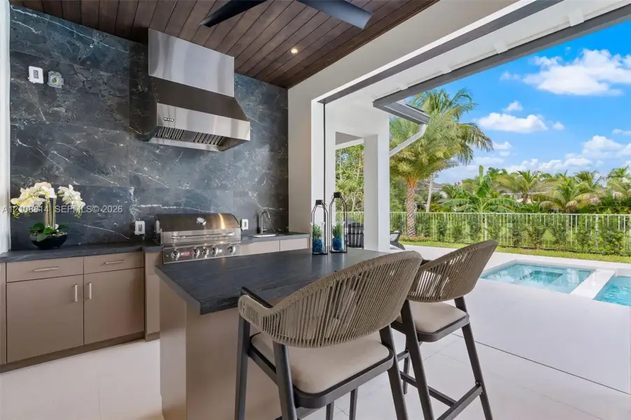 Covered outdoor kitchen featuring a dark stone backsplash, stainless steel range hood, integrated gas grill, flat-panel cabinetry, and dark-toned stone-finish countertop Covered outdoor kitchen featuring a dark stone backsplash, stainless steel range hood, integrated gas grill, flat-panel cabinetry, and dark-toned stone-finish countertop