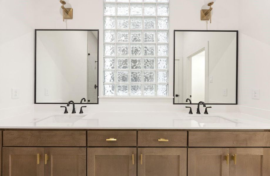 Double vanity detail showcasing warm wood cabinetry, sleek white countertop, black framed mirrors, black faucets, brass-toned sconces, and a glass block window that brings in filtered natural light. The balanced design blends modern contrast with soft warmth for a stylish, spa-like bathroom setting.