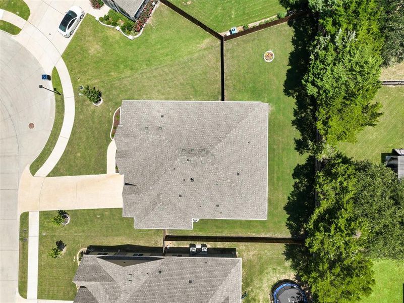 Aerial perspective showcasing a property with a grey shingle roof, extensive green lawn, concrete driveway, and mature trees bordering the rear perimeter Aerial perspective showcasing a property with a grey shingle roof, extensive green lawn, concrete driveway, and mature trees bordering the rear perimeter