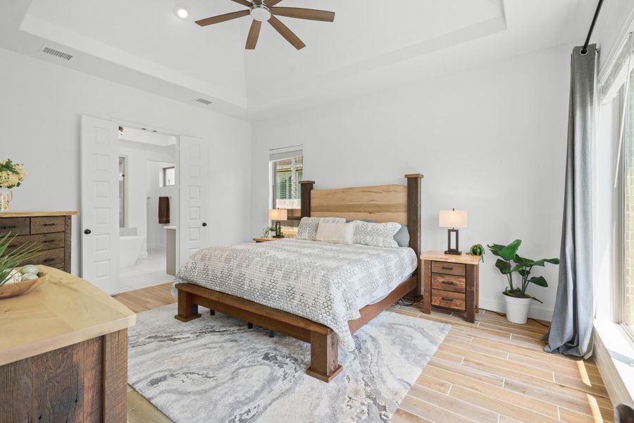 This spacious primary bedroom features a tray ceiling. Natural light flooding in through large windows with light curtains, while a ceiling fan enhances comfort. The room is connected to a primary retreat bathroom through double doors.