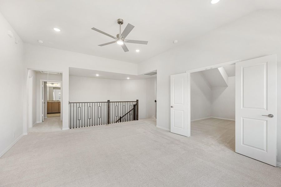 Spacious upstairs landing with ceiling fan, iron stair railing, carpet flooring, and open doors to bathroom and storage room.