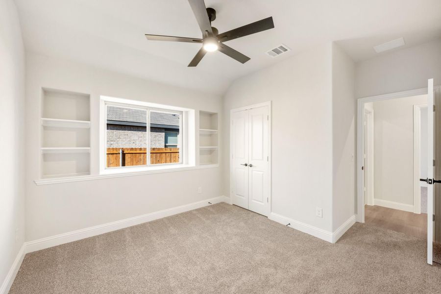 Empty bedroom with carpet flooring, ceiling fan, built-in shelves, and a window with backyard view