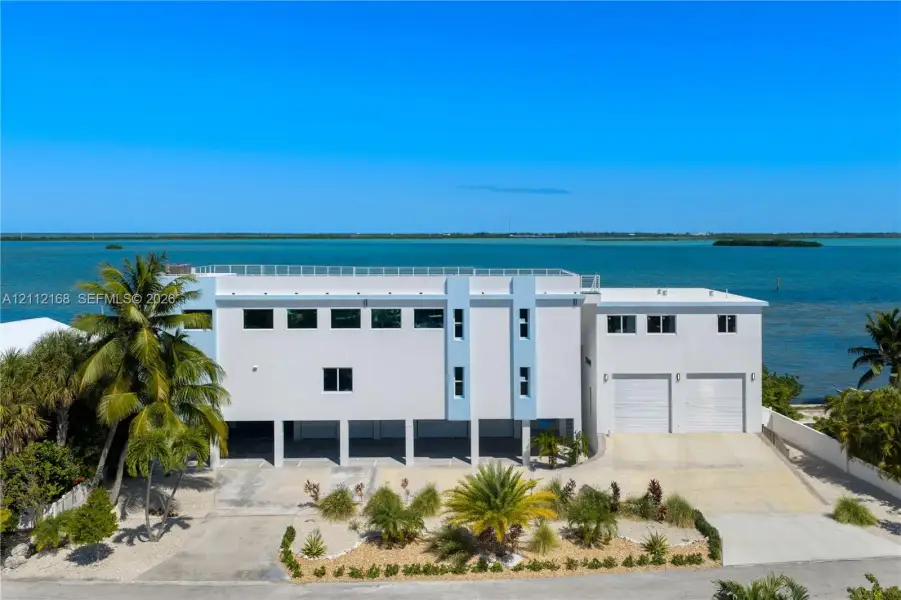 Multi-level coastal residence featuring an elevated structure, rooftop terrace with railing, dual garage bays, expansive concrete driveway, and direct water frontage Multi-level coastal residence featuring an elevated structure, rooftop terrace with railing, dual garage bays, expansive concrete driveway, and direct water frontage