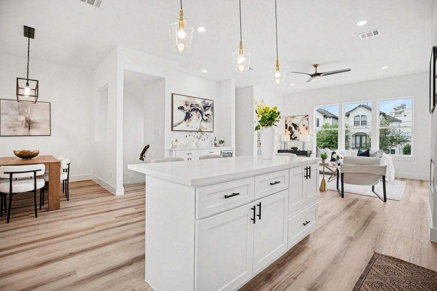 This photo showcases a modern, open-concept living space with a bright kitchen featuring a large island, contemporary light fixtures, and wood flooring. The adjacent dining area and cozy living room are well-lit by large windows, offering a welcoming and airy atmosphere.