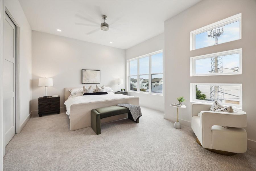 This spacious bedroom features a modern design with neutral tones, large windows for natural light, and a cozy seating area. The room includes a ceiling fan, plush carpet, and sleek furnishings for a comfortable and stylish retreat.