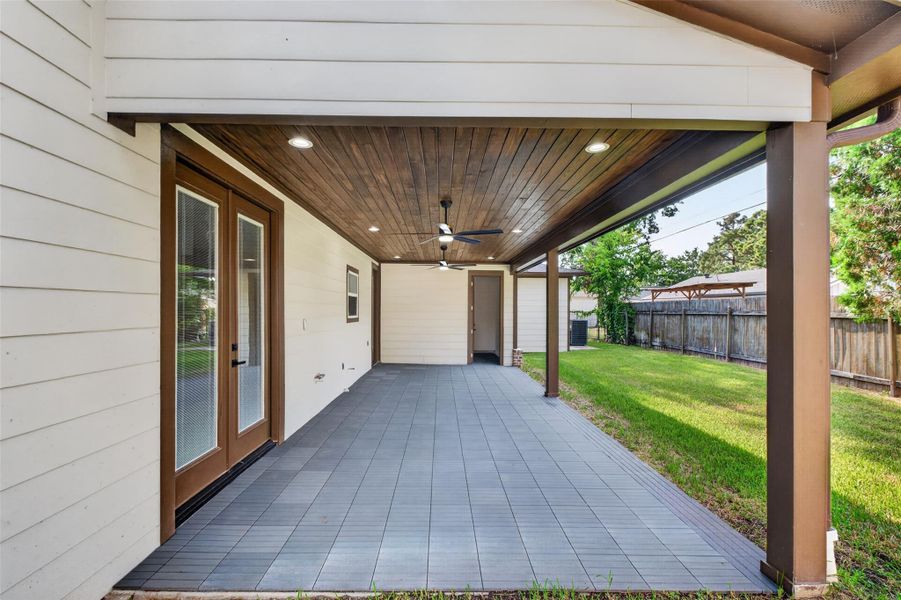 Spacious covered patio with elegant wood ceiling and fan, leading to a lush fenced backyard, perfect for outdoor gatherings.