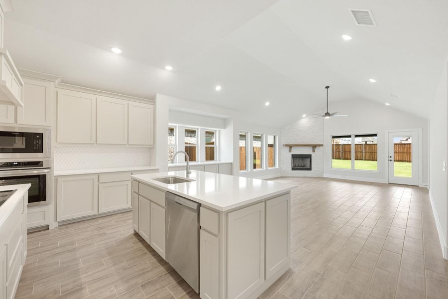 Open kitchen with white cabinets, large island, and stainless appliances opening to living room with brick fireplace