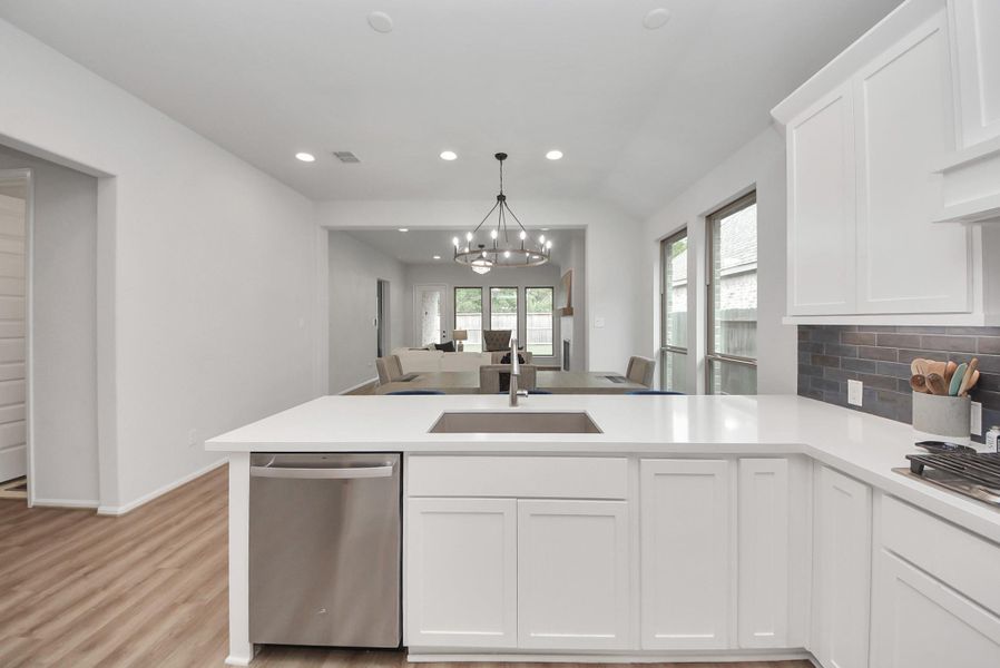 This photo features a modern, open-concept kitchen and living area. The kitchen has sleek white cabinetry, a large island with a sink, and stainless steel appliances. The space flows seamlessly into the living area, which is bright with natural light from multiple windows. The room features wood flooring and a contemporary chandelier, creating a welcoming atmosphere.