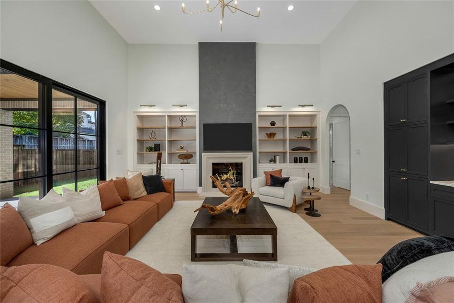 Soaring ceilings define this living space featuring a fireplace with a stone surround, built-in shelving, dark wood-finish cabinetry, and light wood-finish flooring