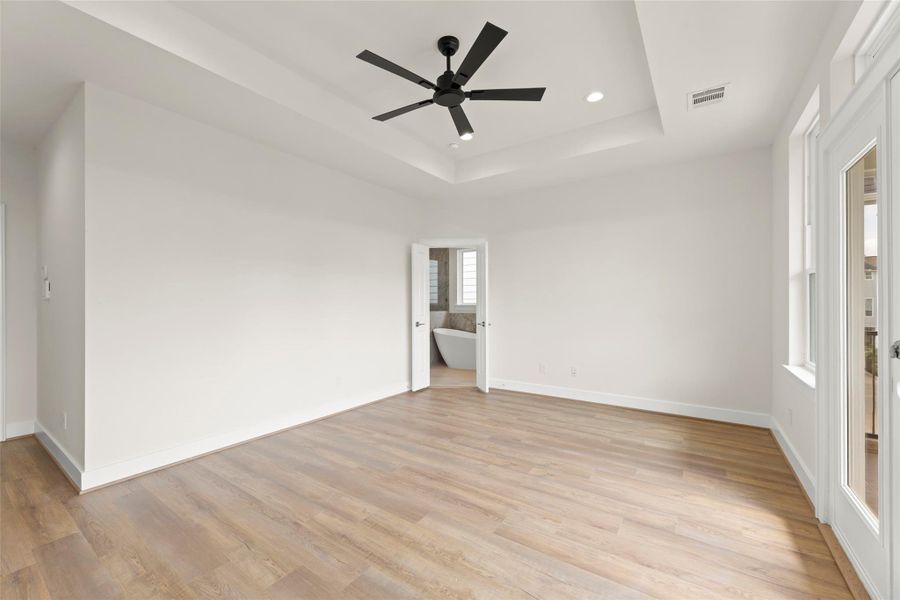 This bright, spacious room features light flooring, a modern ceiling fan, and ample natural light from large windows. A door leads to an en-suite bathroom with a glimpse of a freestanding tub, enhancing luxury living.