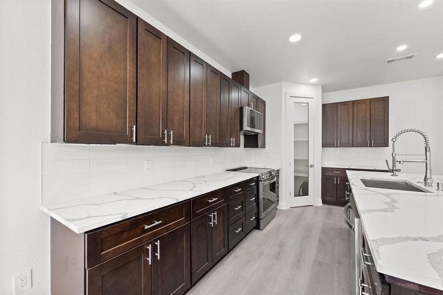 Kitchen with dark wood finish cabinetry, stainless steel appliances, light stone counters, backsplash, and light wood-style flooring