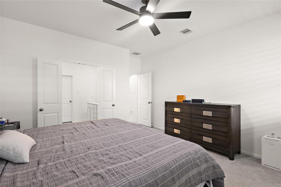 This bedroom features a modern ceiling fan, neutral walls, and a spacious layout. It includes a large bed with a dark wood dresser, and an ensuite bathroom is accessible through a door on the left.