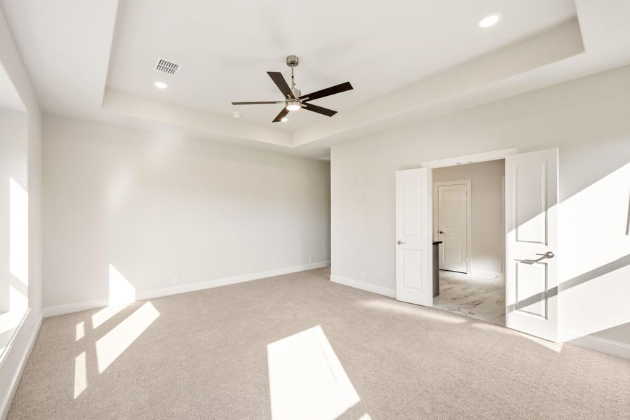 Empty bedroom with tray ceiling, ceiling fan, carpet flooring, and open doors to ensuite bathroom Empty bedroom with tray ceiling, ceiling fan, carpet flooring, and open doors to ensuite bathroom
