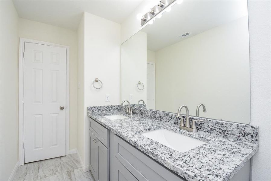 Modern bathroom with double sinks, large mirror, granite countertop, and white door. Bright lighting and neutral color scheme.