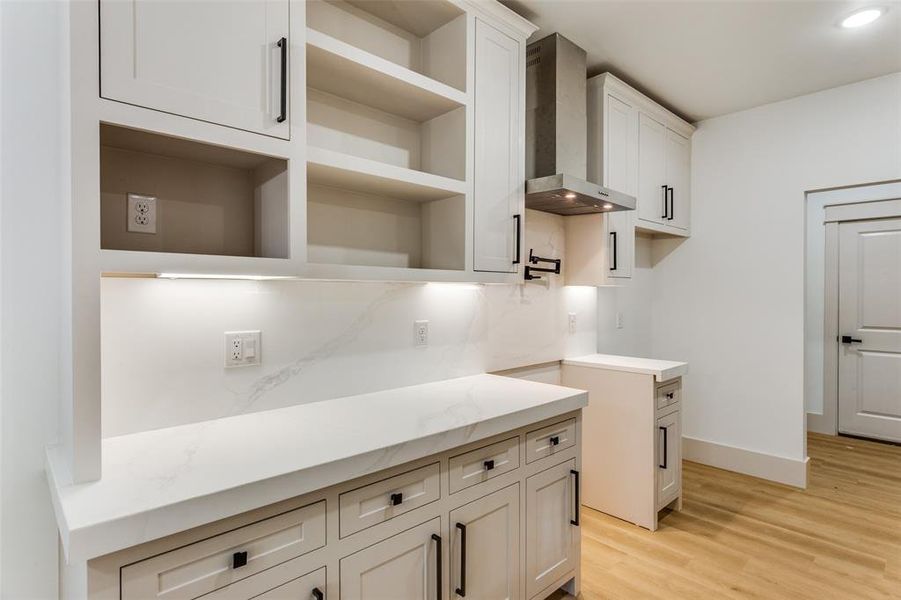 Kitchen with wall chimney exhaust hood, light wood-style floors, light stone countertops, tasteful backsplash, and white cabinets Kitchen with wall chimney exhaust hood, light wood-style floors, light stone countertops, tasteful backsplash, and white cabinets