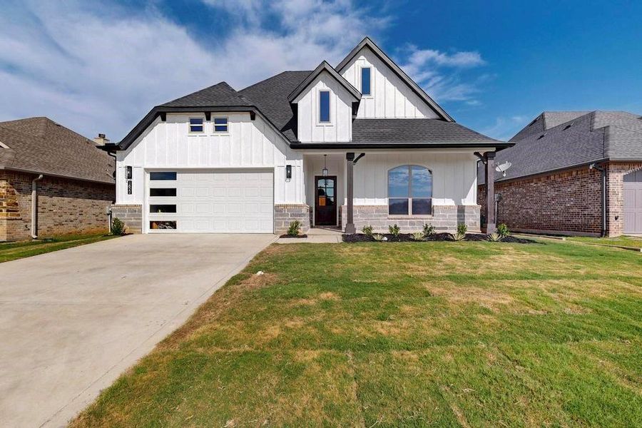 Modern farmhouse style home featuring board and batten siding, a shingled roof, stone siding, and a front lawn