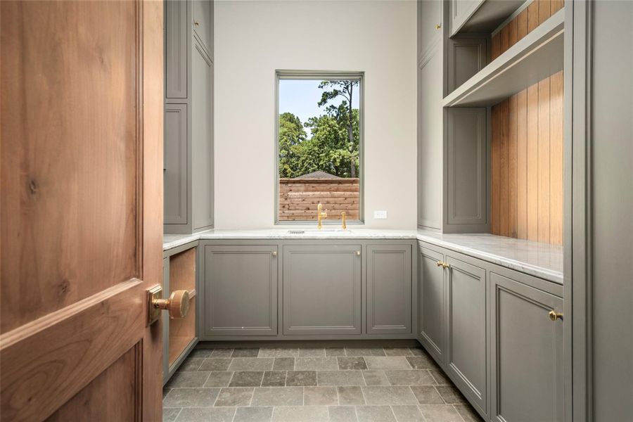 The first-floor laundry room, one of two in the home, combines generous custom cabinetry with expansive stone countertops, open shelving and a dedicated utility sink. Warm wood detailing and brass accents elevate this highly functional space, while a large window brings in natural light. The first-floor laundry room, one of two in the home, combines generous custom cabinetry with expansive stone countertops, open shelving and a dedicated utility sink. Warm wood detailing and brass accents elevate this highly functional space, while a large window brings in natural light.