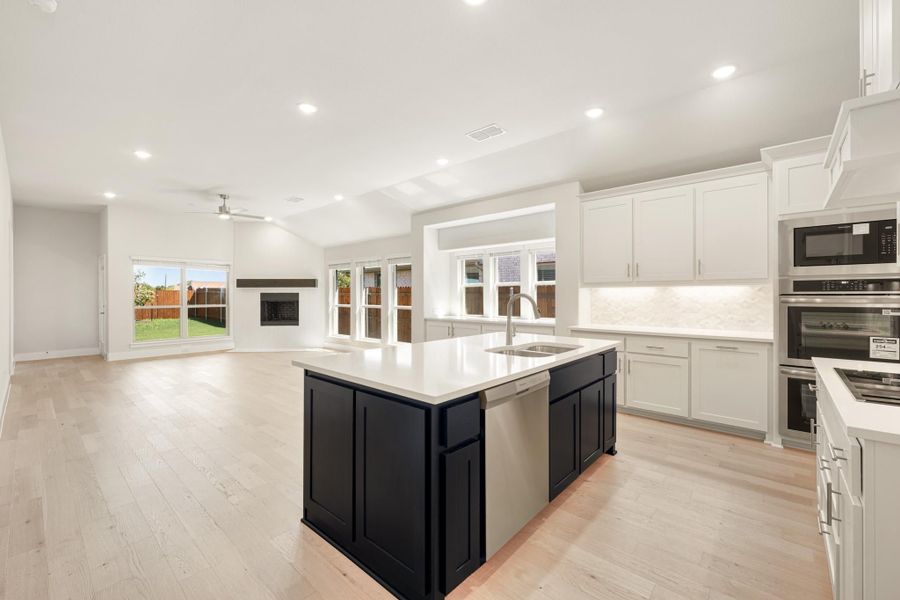 Open kitchen with dark island, white cabinets, stainless appliances, and light wood floors open to living area with fireplace.