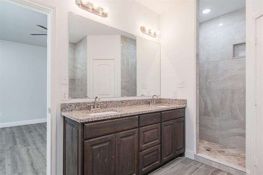 Full bath with double vanity, baseboards, tiled shower, wood finished floors, and a sink