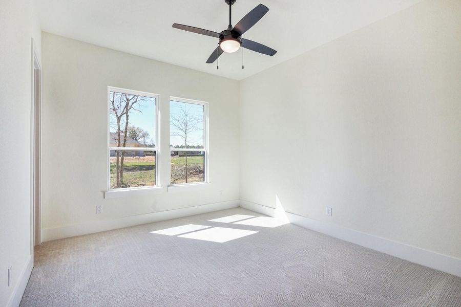 This bright bedroom features light-colored walls and carpet, with two large windows offering plenty of natural light. A modern ceiling fan is centrally placed, providing both style and functionality.
