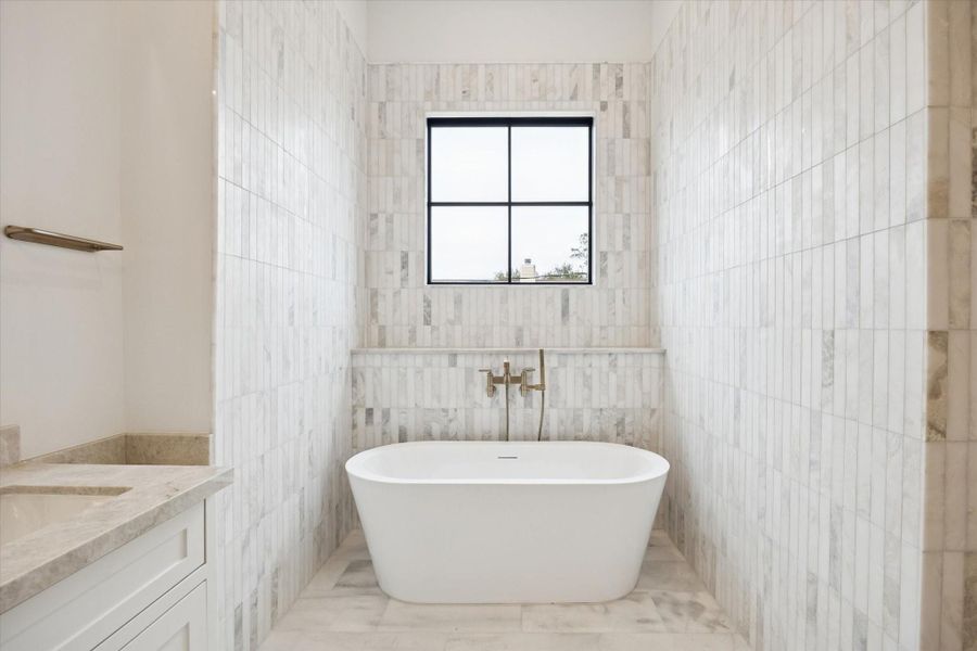 Freestanding soaking tub framed by full height marble tile and a black framed picture window creates a true spa retreat. Designer brass fixtures and seamless stone flooring elevate the space with timeless elegance and refined detail.