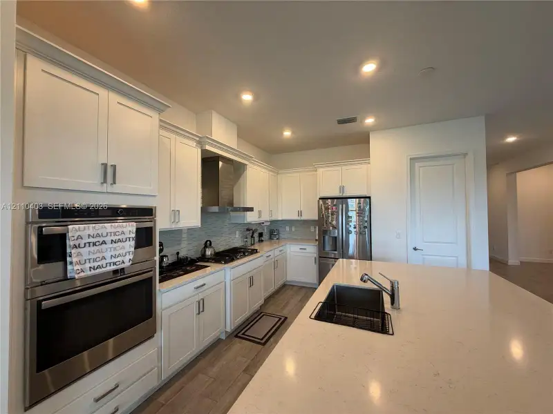 Kitchen featuring white shaker-style cabinetry, stainless steel double wall ovens, a gas cooktop with a range hood, quartz countertops, and a center island with an undermount sink